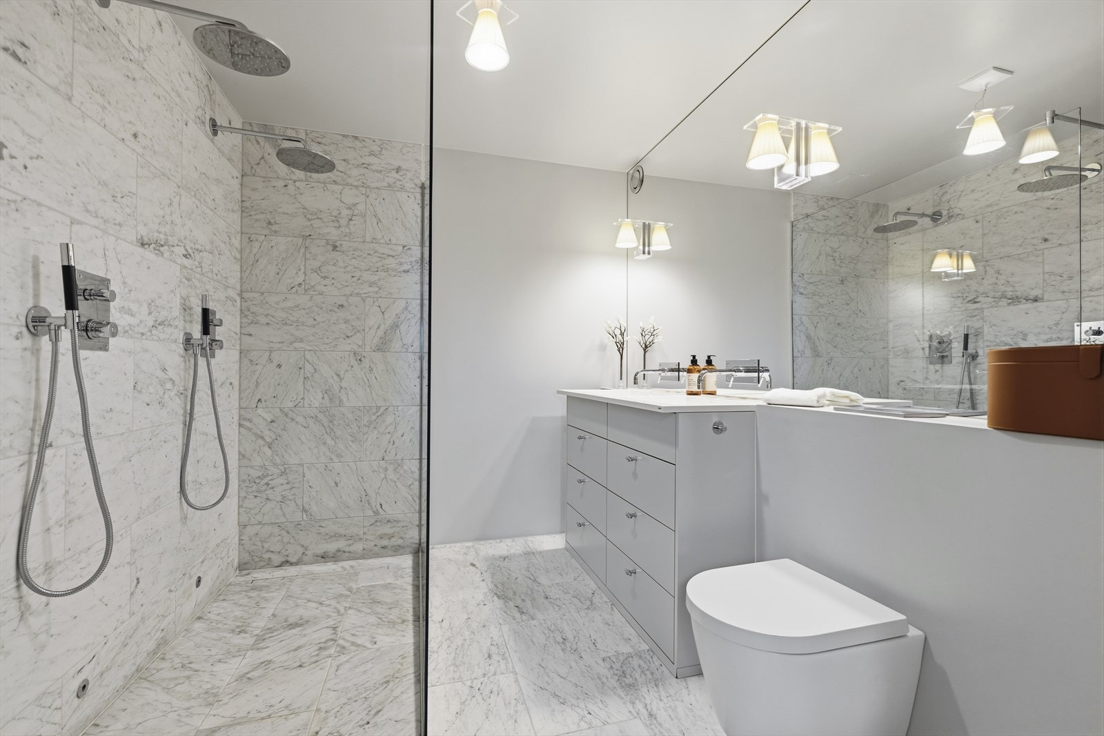 Badet inneholder veggmontert toalett, stor dusjnisje med to dusjhoder og glassvegg, sluk i dusj, to servanter med blandebatterier og stort speil på vegg. Carraramarmor fliser fra Stenprosjekt Galleribilde