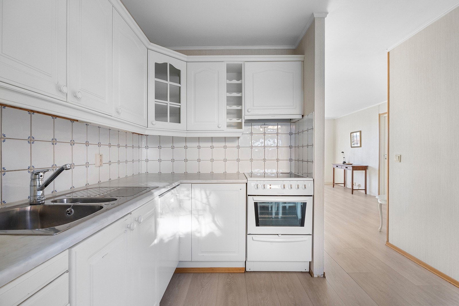 Kjøkkeninnredning med profilerte lakkerte skapfronter, laminert benkeplate med fliser på bakvegg, 1 1/2 oppvaskkum med benkebeslag, ventilator og frittstående hvitevarer som komfyr, oppvaskmaskin, kjøl/- fryseskap Galleribilde