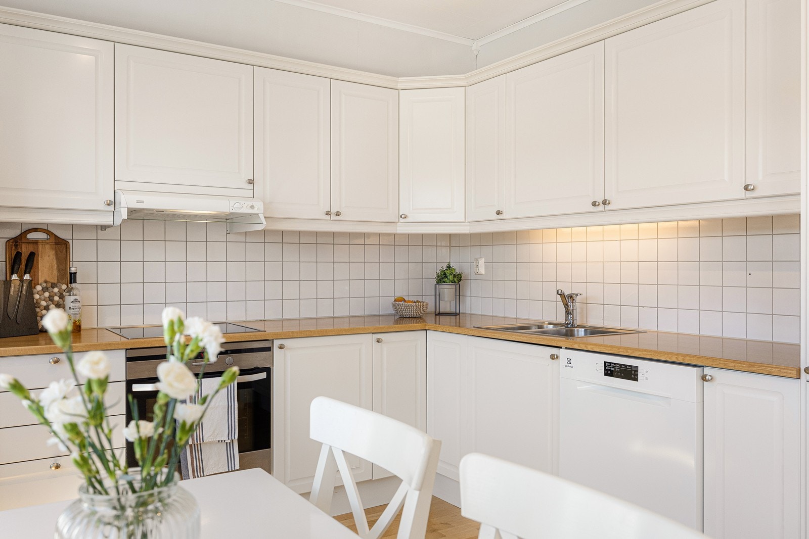 Kjøkkenet er oppført i en funksjonell L-form med god skap- og benkeplass. Innredningen har hvite, profilerte fronter, dobbel oppvaskkum og en laminert benkeplate i gyllen farge. Galleribilde