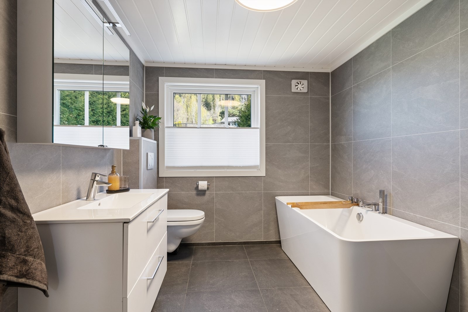 Badet er oppusset nå i 2026. Det er store fliser på både vegger og gulv. Servant og speilskap, vegghengt toalett og badekar. Galleribilde