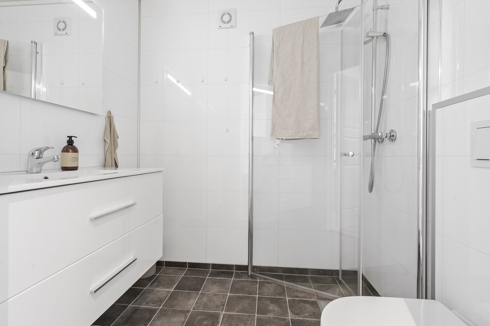 Baderom fra byggeår (2022) med keramiske fliser på gulv, baderomsplater på vegger og malt panelhimling med integrerte spotlights. Badet inneholder servant med møblement og speil, vegghengt toalett og dusjløsning med dusjgarnityr. Galleribilde