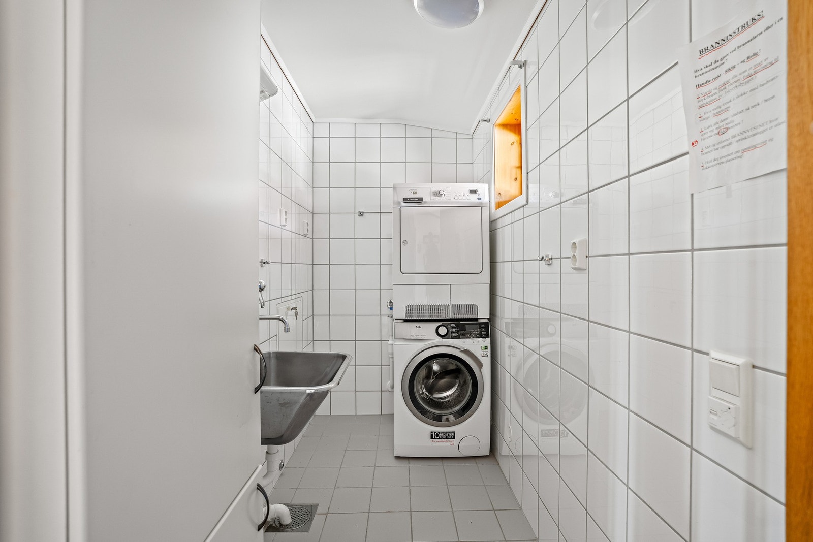 Praktisk flislagt vaskerom med varmekabler i gulvet. Innredning bestående av veggskap med glatte fronter, skyllekum i rustfritt stål og opplegg for vaskemaskin. Galleribilde