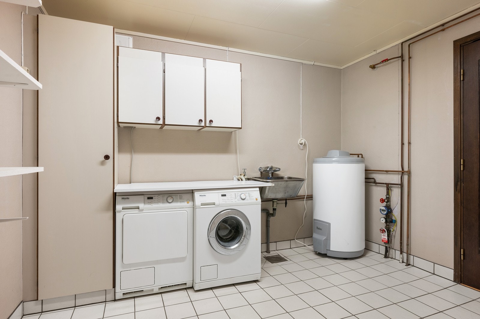 Vaskerom i underetasjen har flislagt gulv og malte vegger. Det er innredet med skapfronter, utslagsvask og opplegg for vaskemaskin. Rommet er ventilert via en loftsvifte, og varmtvannstanken er plassert her. Galleribilde