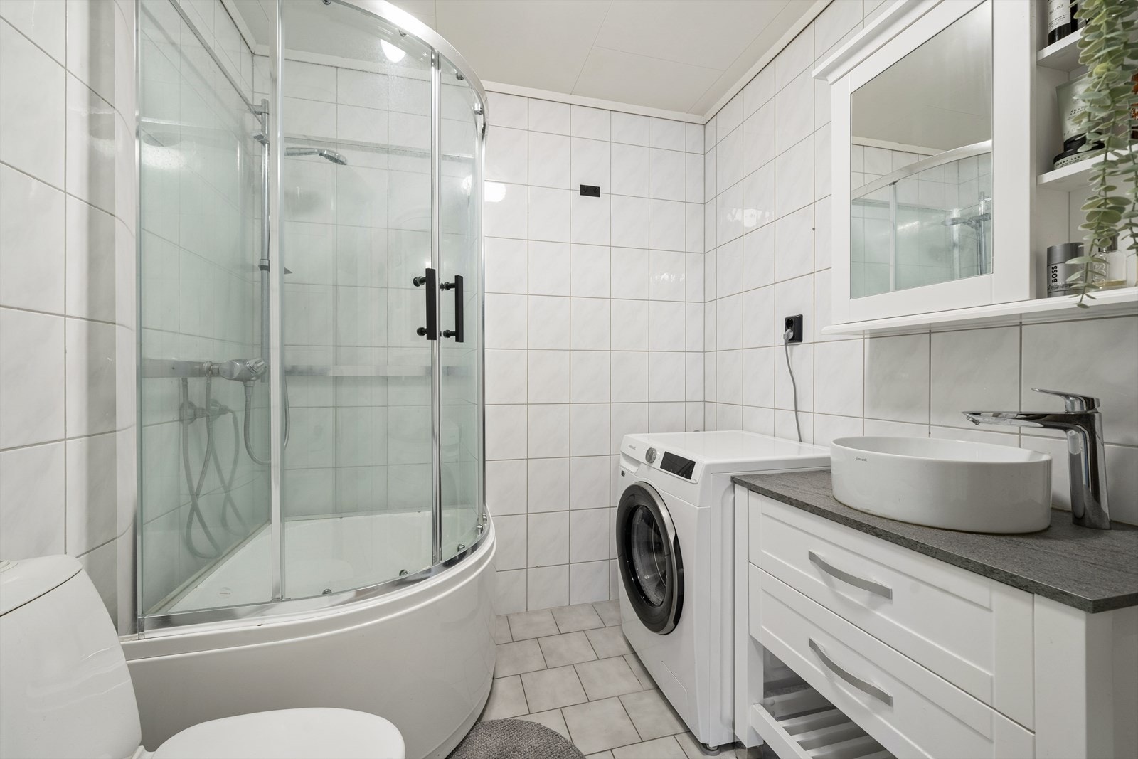 Klassisk baderom med tidløse fliser på gulv og vegger. Badet er utstyrt med dusjkabinett som kan fungere som badekar, baderomsinnredning med servant og speilskap samt opplegg for vaskemaskin. Galleribilde