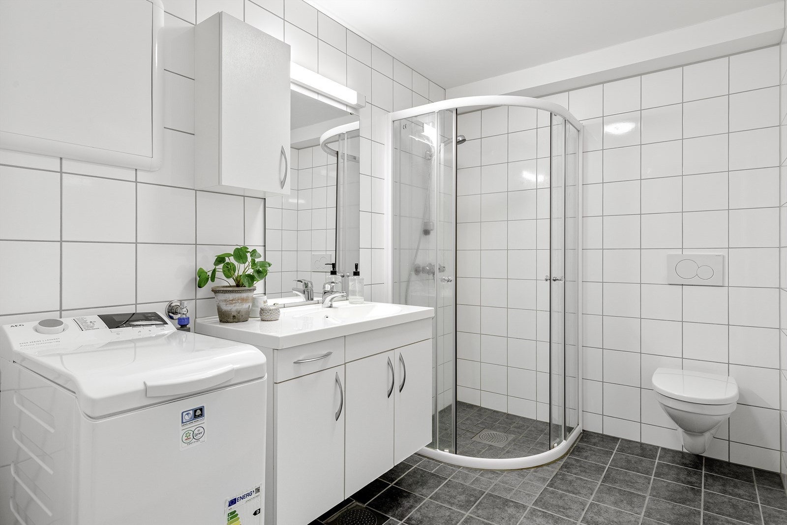 Badet er utstyrt med dusjhjørne med skøyvedører, nedfelt servant med underskåp, spegel med lyslist, sideskåp, vegghengt toalett og opplegg for vaskemaskin. Galleribilde