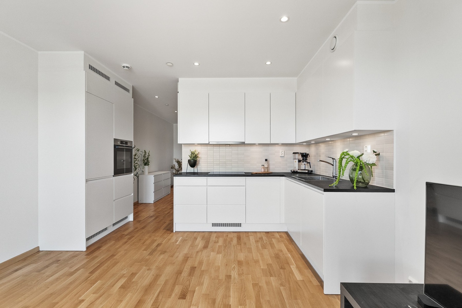 Tidløst og pent kjøkken som er godt utbygget. Hvite slette fronter og benkeplate av laminat.Fliser på vegg over benk. Downlights under overskap. Galleribilde