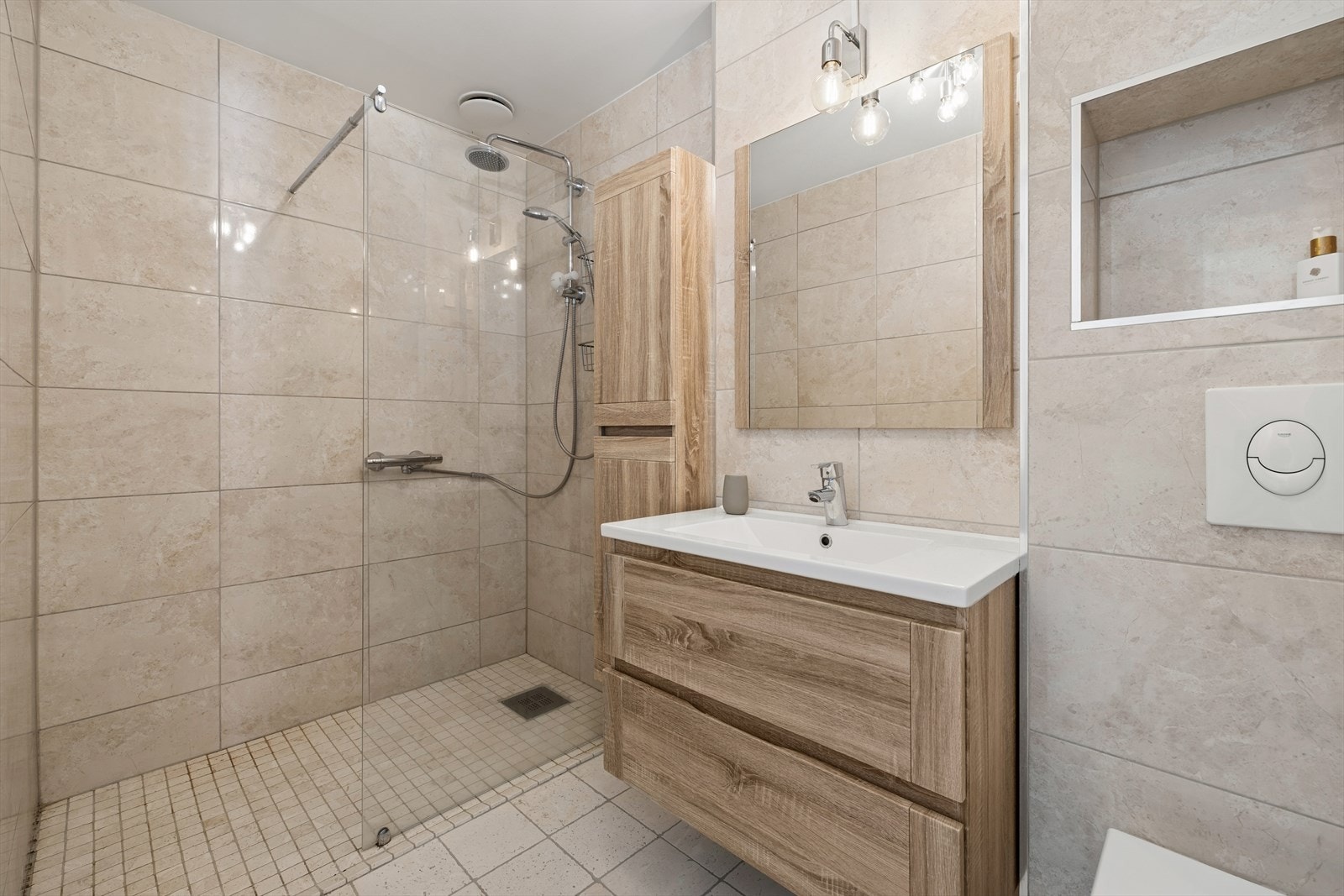 Badet er flislagt. Rommet har varmekabler. Det er innredning med nedfelt servant, veggmontert toalett og dusjvegger/hjørne. Galleribilde