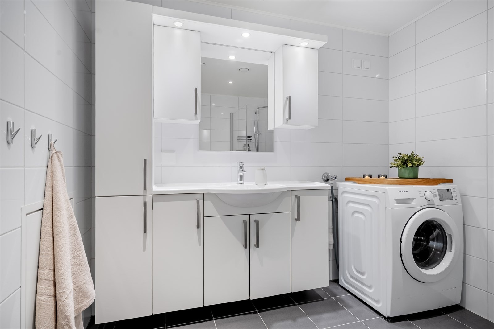 Badet inneholder heldekkende servant over innredning, speil med lys, høyskap, vegghengt toalett og dusj med glassvegger - Opplegg for vaskemaskin Galleribilde
