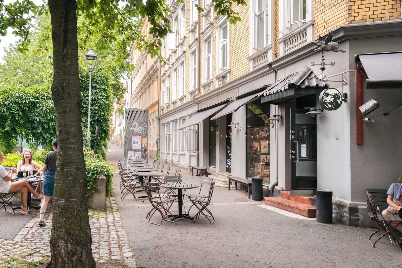 Området byr på rikelig med uteservering med flere bord og stoler langs en bygård i et urbant område. Galleribilde