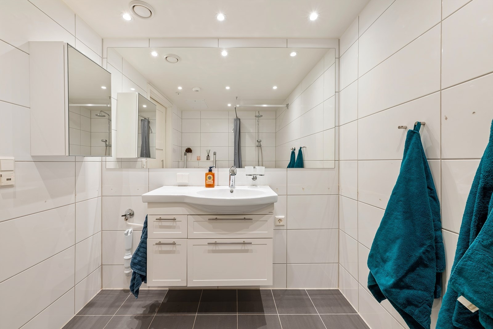 Badet er utstyrt med vegghengt toalett, servant med underskap og speil, samt dusjløsning med dusjarmatur og forheng. Fliser på gulv og vegger samt varmekabler i gulv sikrer et behagelig og lettstelt baderom. Galleribilde