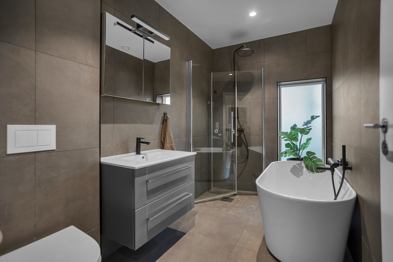 Badet har et moderne uttrykk med flislagte overflater, dusjhjørne og frittstående badekar. Vindusflater bidrar med naturlig lysinnslipp og en luftig romfølelse. Galleribilde