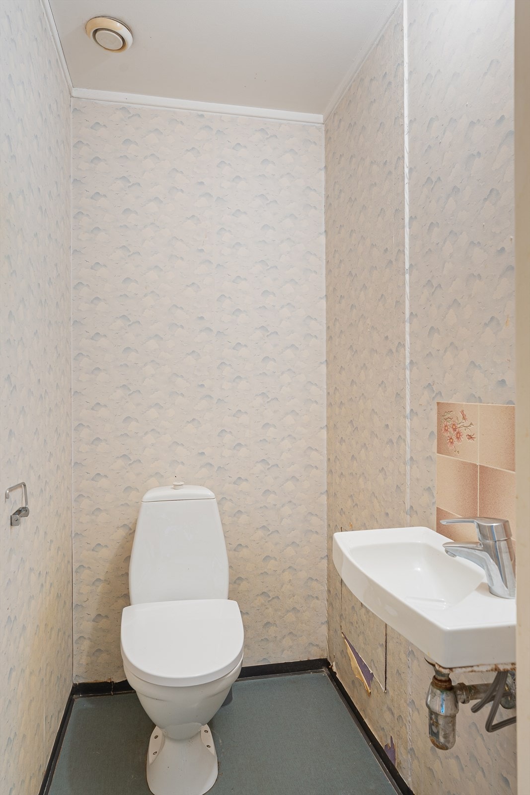 Separat, eldre WC-rom med renoveringsbehov. Belegg på gulv. Tapetserte overflater på vegger og malte tak. Det er installert gulvmontert toalett og vask med blander. Galleribilde
