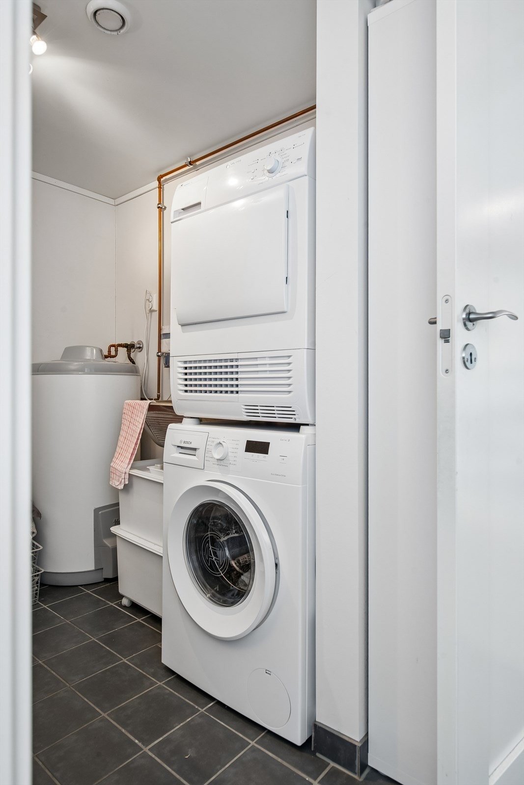 Praktisk vaskerom i føreste etasje med fliser på gulvet, gulvvarme og opplegg for vaskemaskin. Her er også boligens hovedstoppekran plassert. Galleribilde