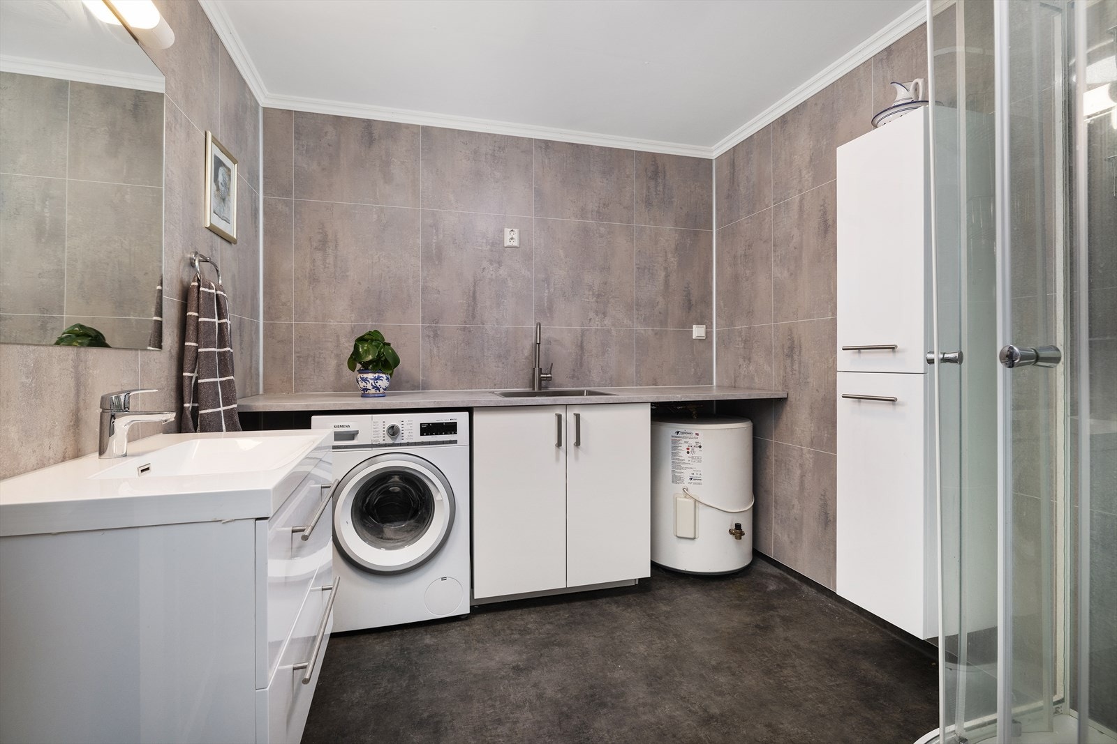 Badet er utstyrt med servantinnredning, benkeinnredning med kum, underskap og nisje for vaskemaskin og varmtvannsbereder, vegghengt skap og dusjkabinett. Galleribilde