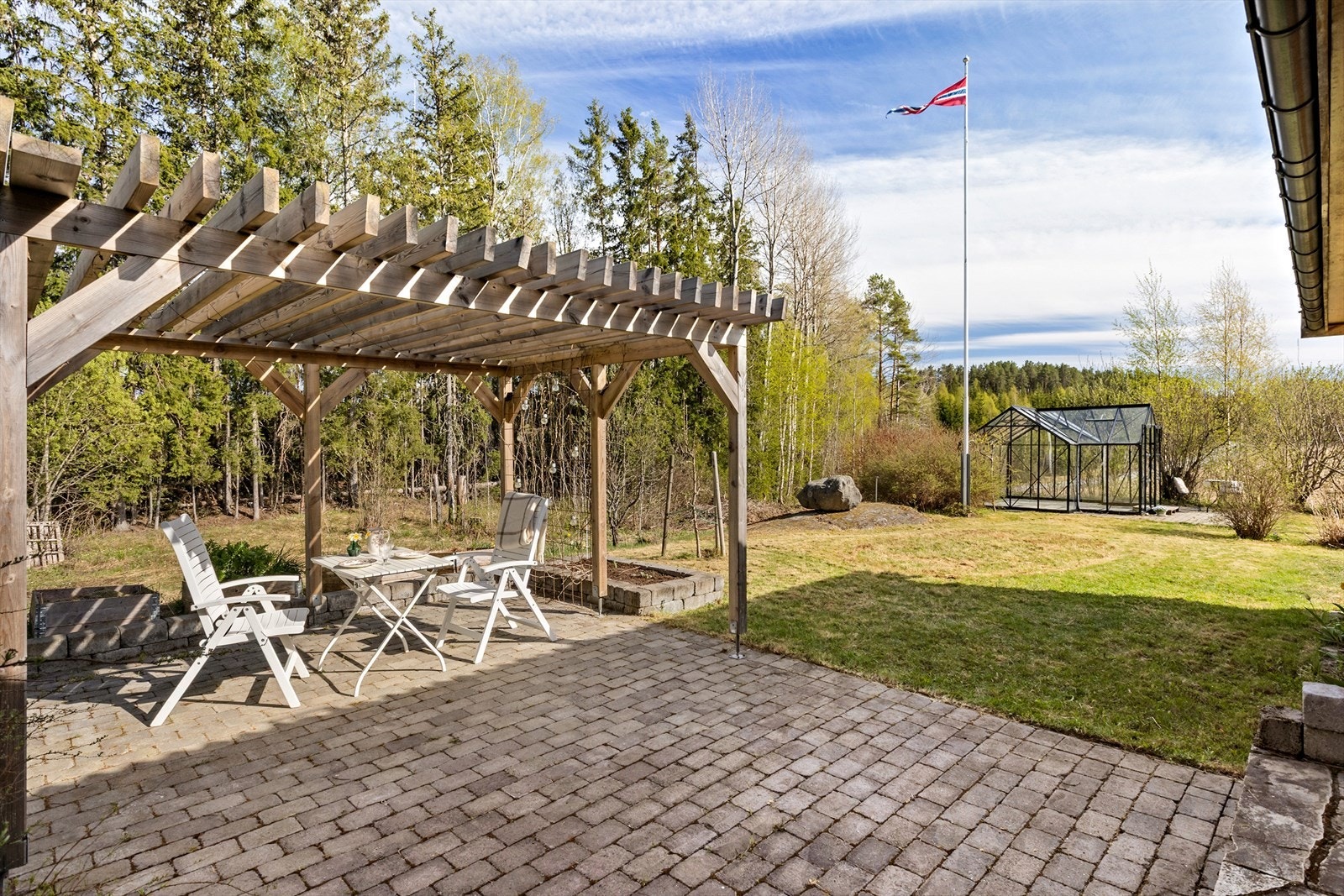 Hellebelagt uteområde, pergola, gressplen med busker og beplantning, flaggstang og drivhus. Galleribilde