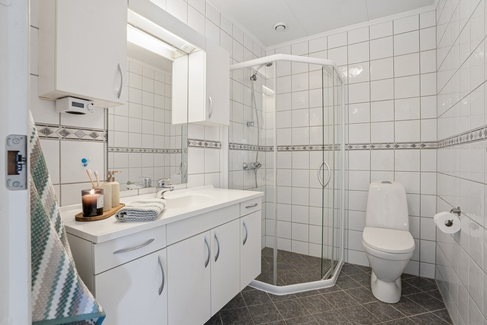 Badet er innredet med hvit baderomsinnredning med heldekkende servantplate, speil og overskap, samt gulvmontert toalett og dusjhjørne. Galleribilde
