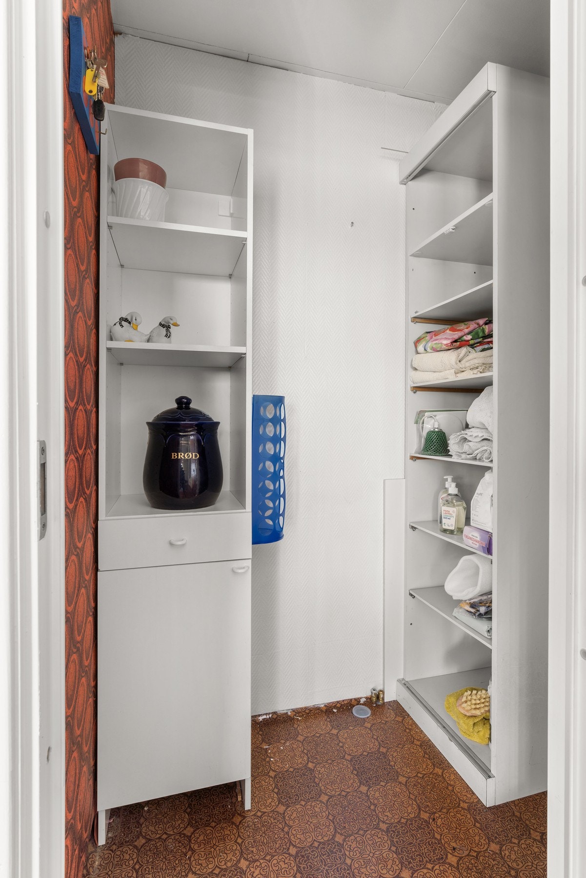Rommet har hvite hyller og skap, samt brunt gulvbelegg med mønster. Veggene er delvis tapetsert. Galleribilde