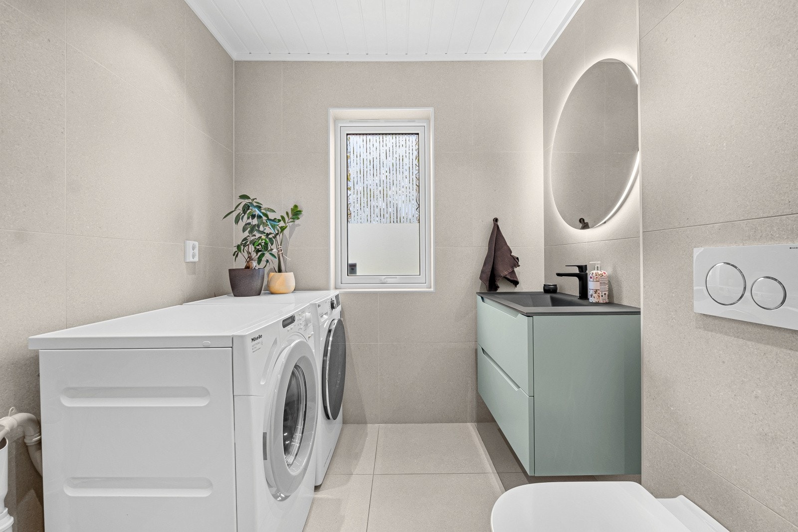 Badet har en moderne baderomsinnredning med nedfelt servant over skuffeseksjon, samt speil med integrert belysning over. Videre er det vegghengt toalett, dusjhjørne og opplegg for vaskemaskin. Galleribilde