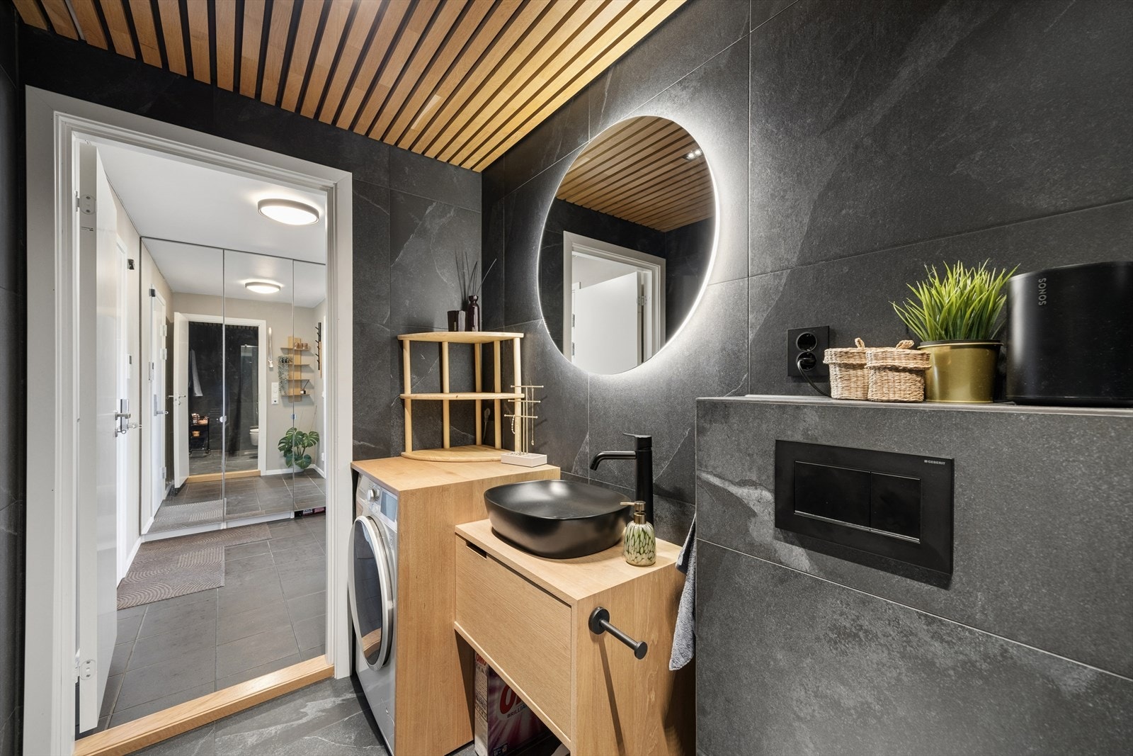 Badet har et moderne uttrykk med mørke fliser og detaljer i lyst treverk som gir en lun og behagelig atmosfære. Det runde speilet med integrert belysning og spiletaket tilfører rommet fine detaljer og et helhetlig preg. Galleribilde