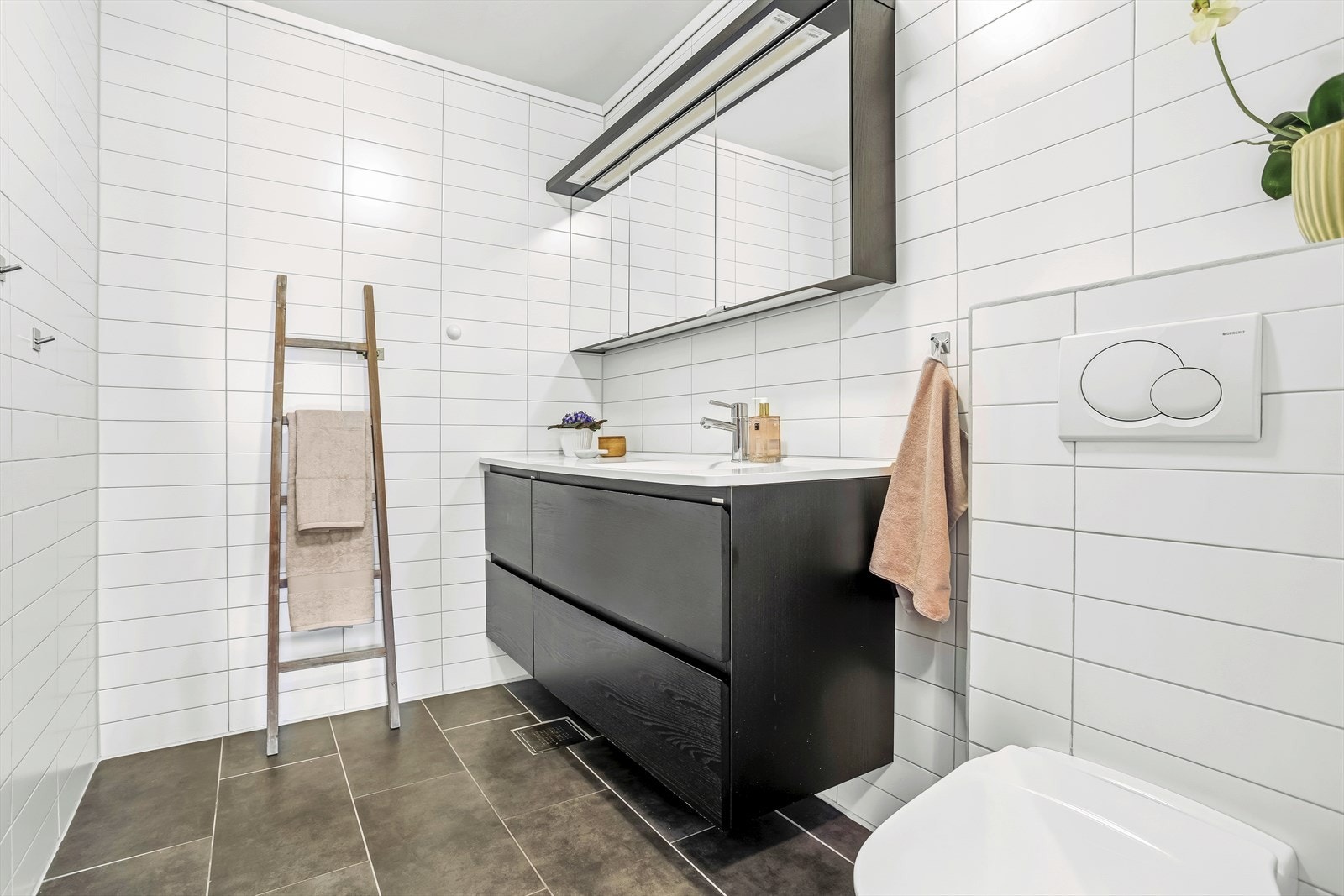 Bad med fliser på gulv og vegger, utstyrt med servant, vegghengt toalett, dusj og varmekabler, samt vaskerom med tapet på vegger, belegg på gulv, skyllekar og opplegg for vaskemaskin. Galleribilde