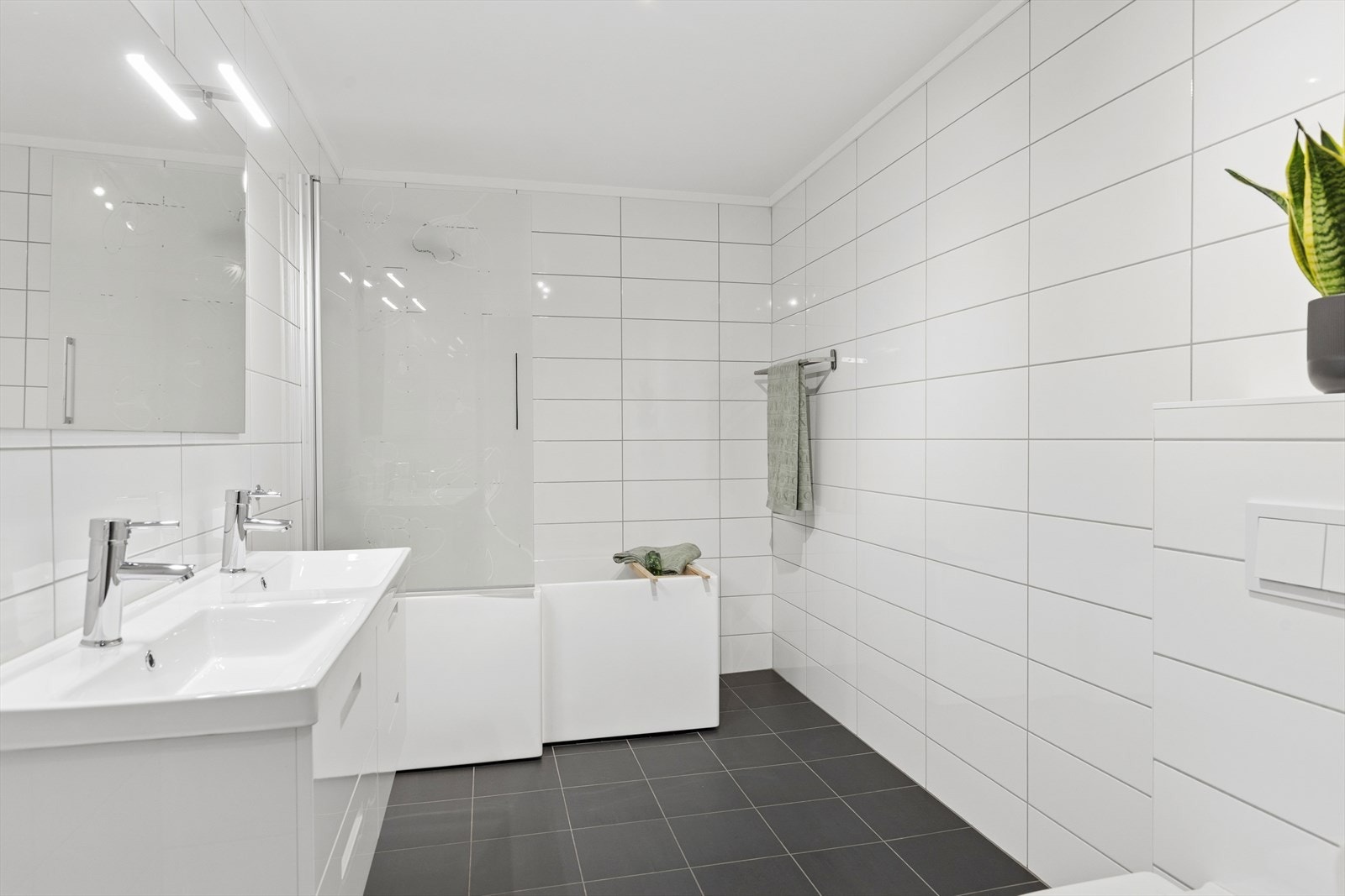 Lekkert bad med fliser. Rommet har baderomsinnredning med nedfelt dobbel servant, badekar med dusjvegg og veggmontert toalett. Galleribilde