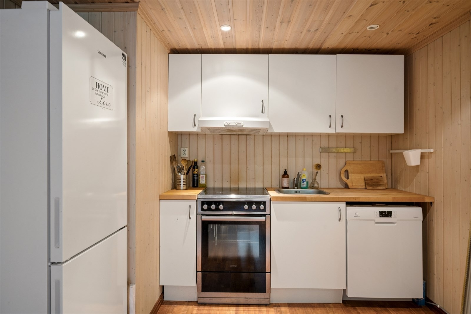 Kjøkkeninnredning med lyse glatte fronter, benkeplate av heltre og belysning under overskap. Det er kjøkkenventilator med avtrekk ut og opplegg for vaskemaskin. Galleribilde