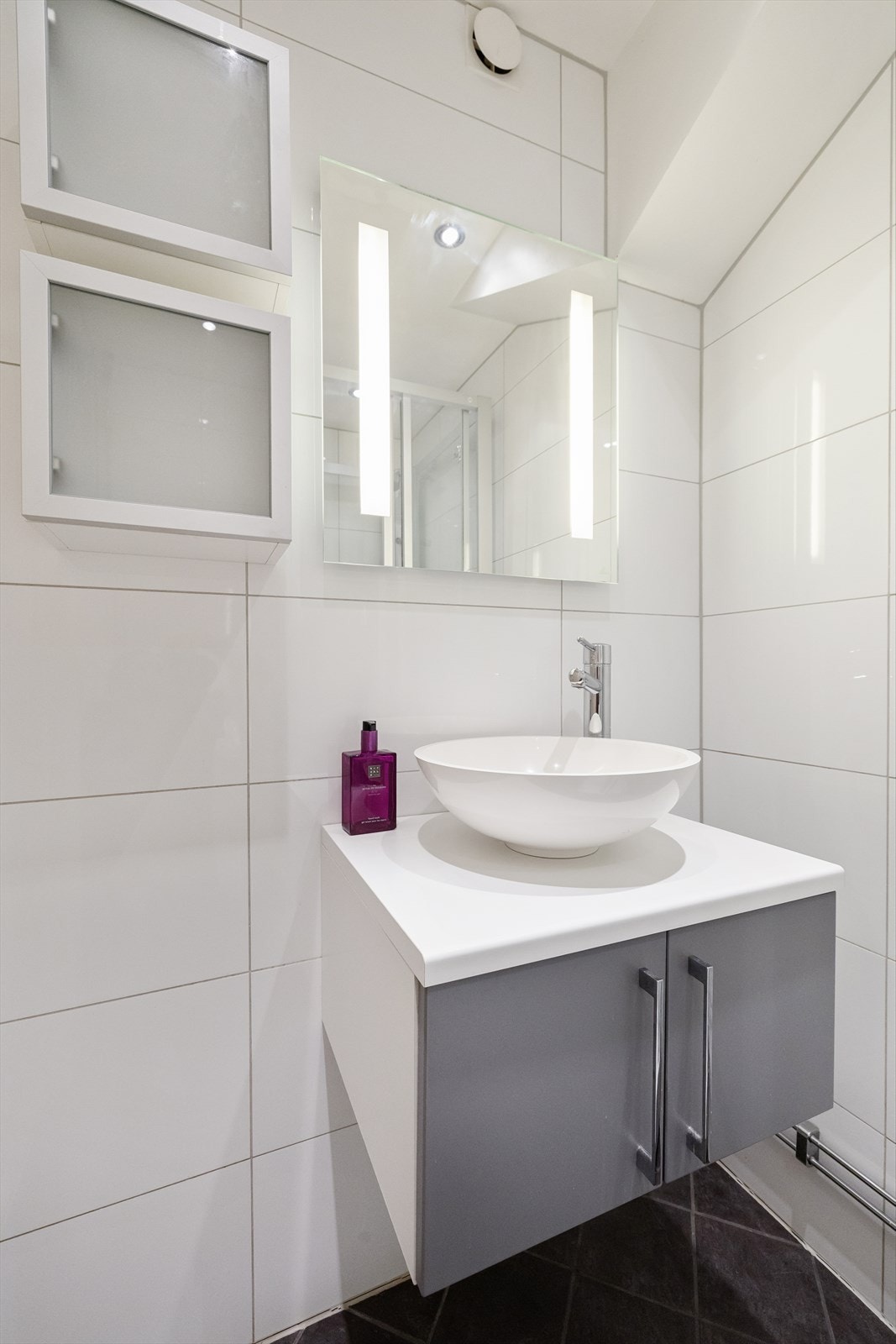 Det er flislagt gulv med termostatstyrte varmekabler, baderomsinnredning med glatte folierte fronter, servantbolle plassert på laminat benkeplate, vaskemaskin (smal modell), dusjkabinett med skyvedører, sluk i sone og dusjutstyr. Galleribilde