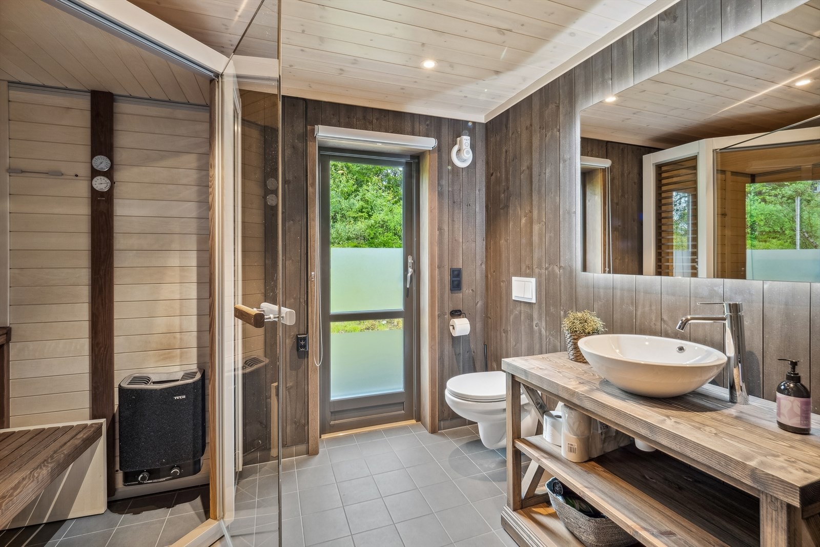 Det andre baderommet har brunbeiset panel på veggene, grå fliser i dusjsonene og grå fliser på gulvet både på badet og i badstuen. Badet blir oppvarmet med elektriske varmekabler i gulvet. Galleribilde