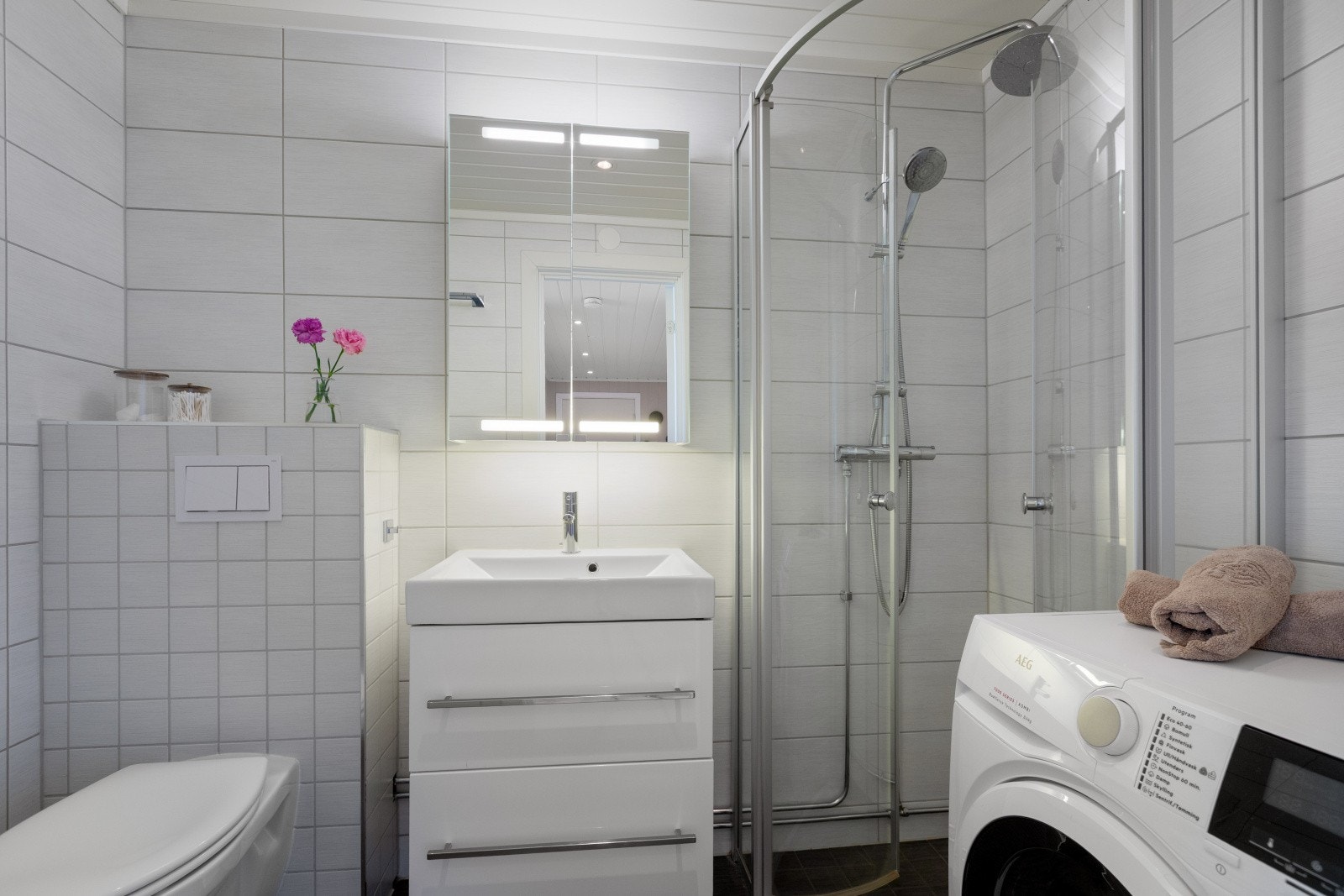 Badet er pent innredet med mørkegrå fliser på gulv og hvite veggfliser. Det er opplegg for vaskemaskin og tørketrommel på badet. Galleribilde
