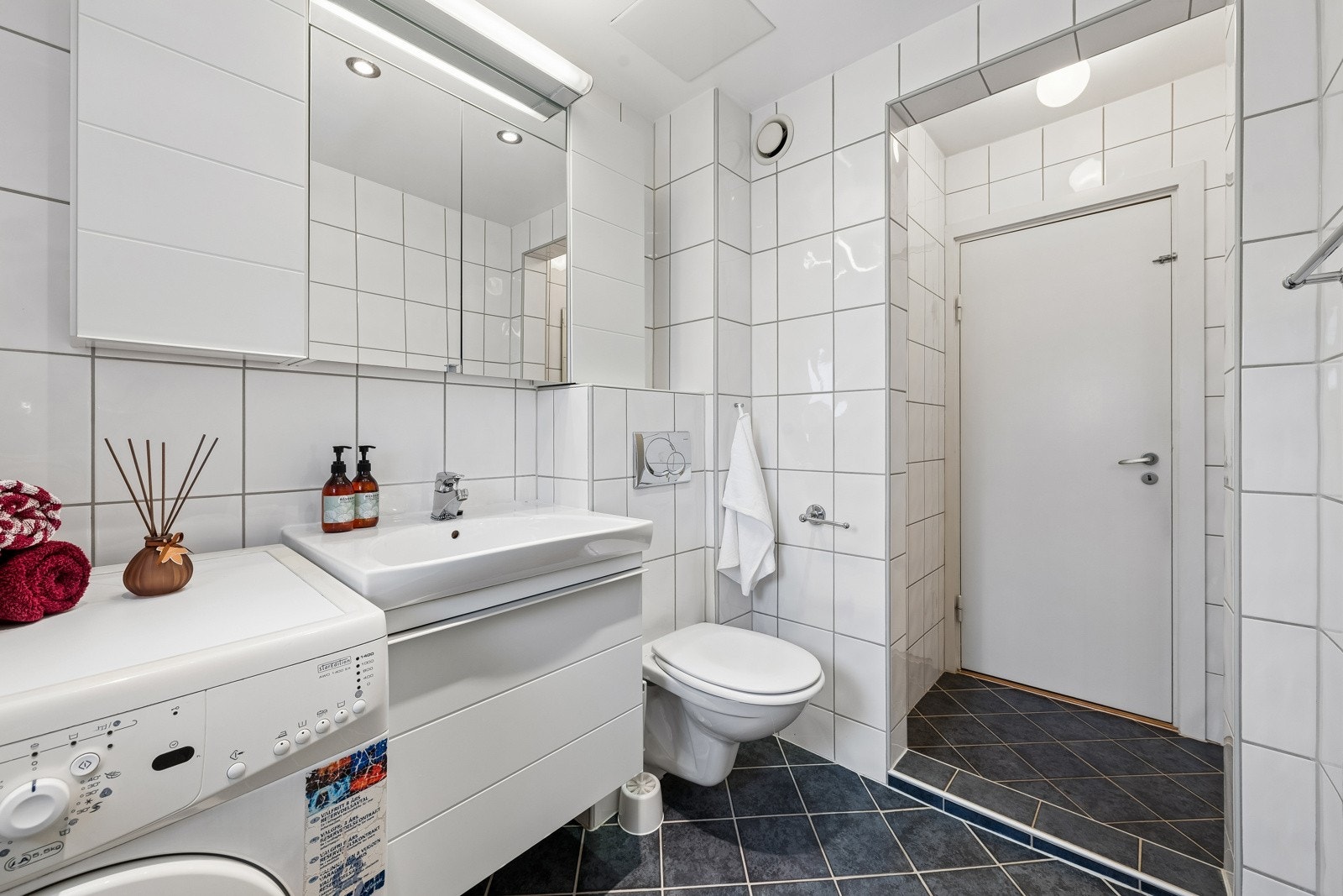 Badet er utstyrt med åpen dusj, servant med underskap, speilskap med lys over og veggmontert klosett. Opplegg for vaskemaskin. Galleribilde