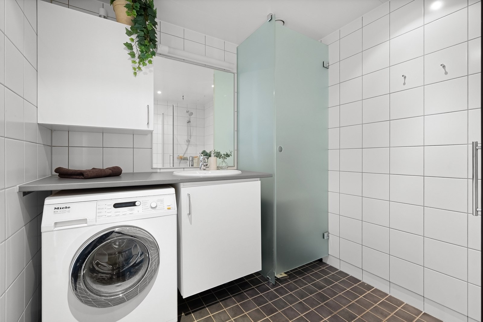 Badet er innredet med vegghengt toalett, dusjhjørne med svingbare glassdører og baderomsmøbel med skap, nedfelt servant og speil. Galleribilde