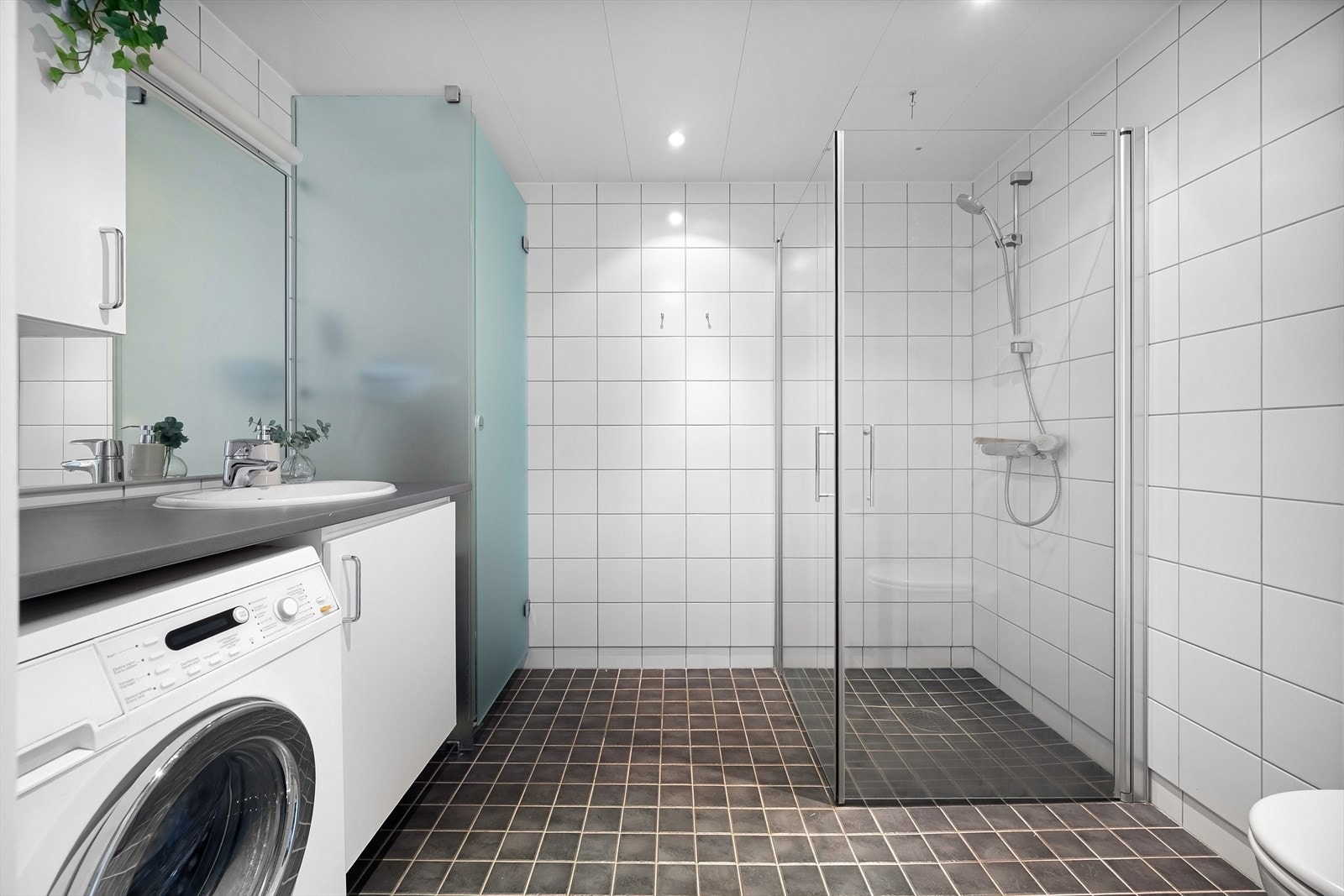 Stort, tidløst bad med flislagte overflater, elektrisk gulvvarme og lakkerte stålplater i himlingen med innfelte downlights. Det er opplegg for vaskemaskin. Galleribilde