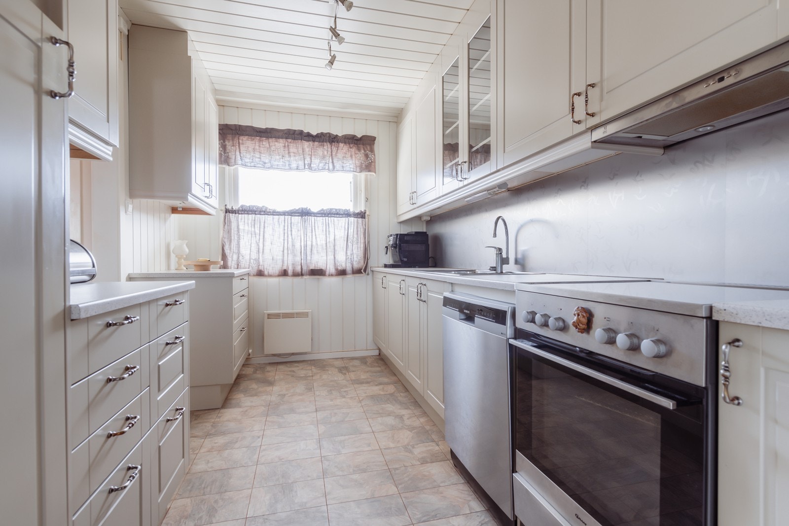 Kjøkkeninnredning med malte fronter, laminat benkeplate, stål oppvaskkum, mekanisk ventilator og frittstående hvitevarer. Galleribilde