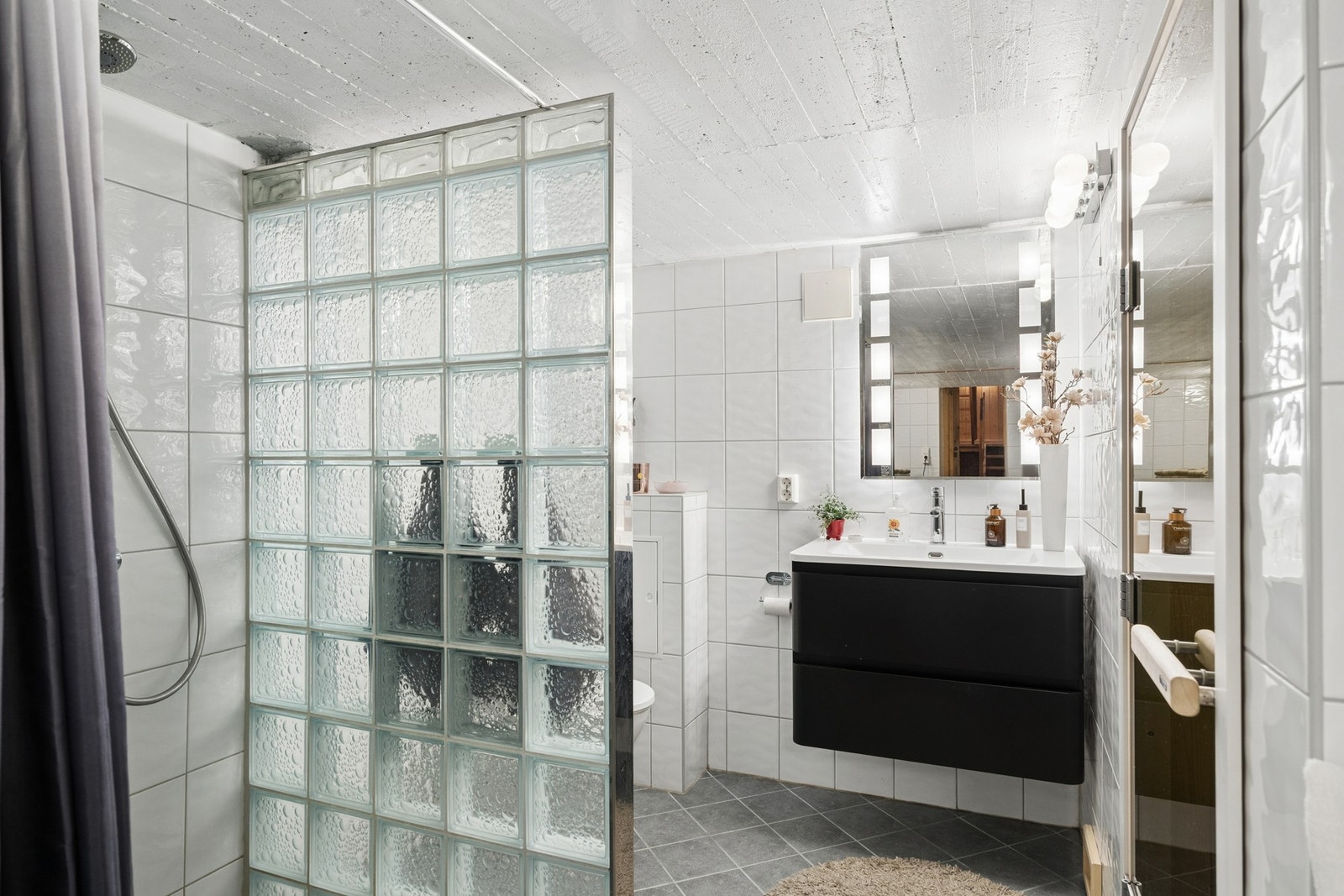 Baderommet har fliser på gulv og vegger, sort servantinnredning med skuffeseksjon, dusjnisje med sprutvegger av glassbyggerstein, opplegg for vaskemaskin og gulvstående toalett. Galleribilde
