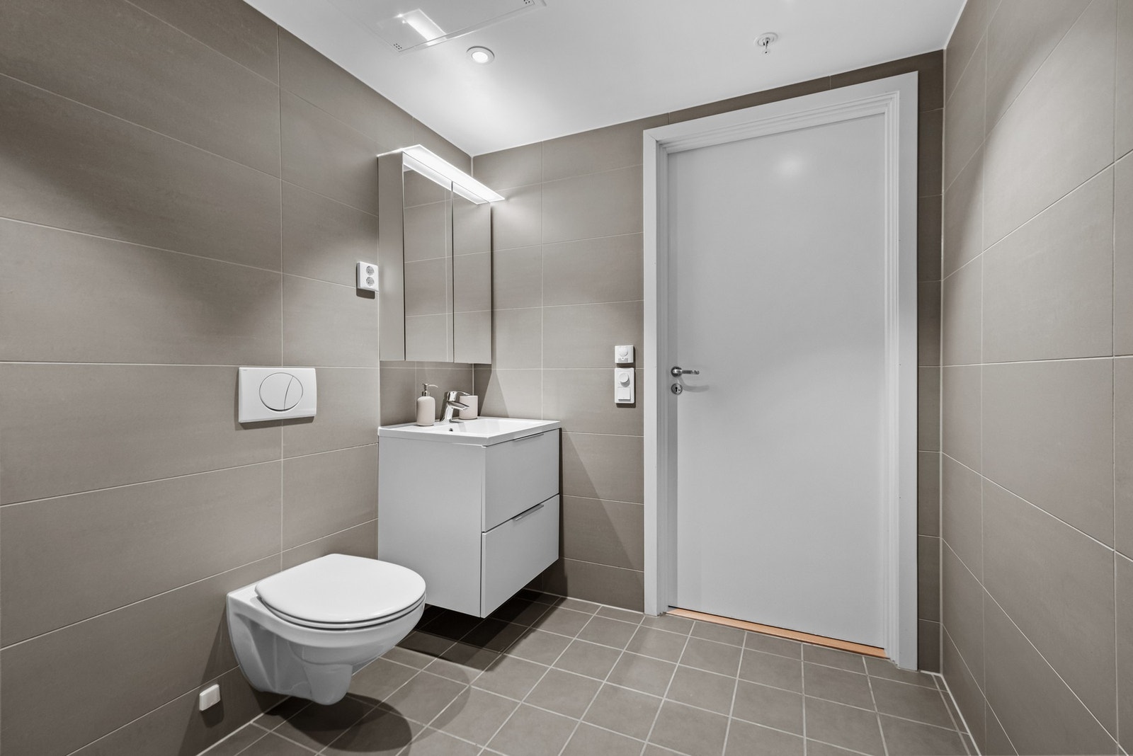 Badet er videre utstyrt med nedsenket himling med downlights, vegghengt hvit servant med skuffer og overliggende servant med ett-greps armatur, delikate fliser på gulv og vegger og vannbåren gulvvarme. Galleribilde