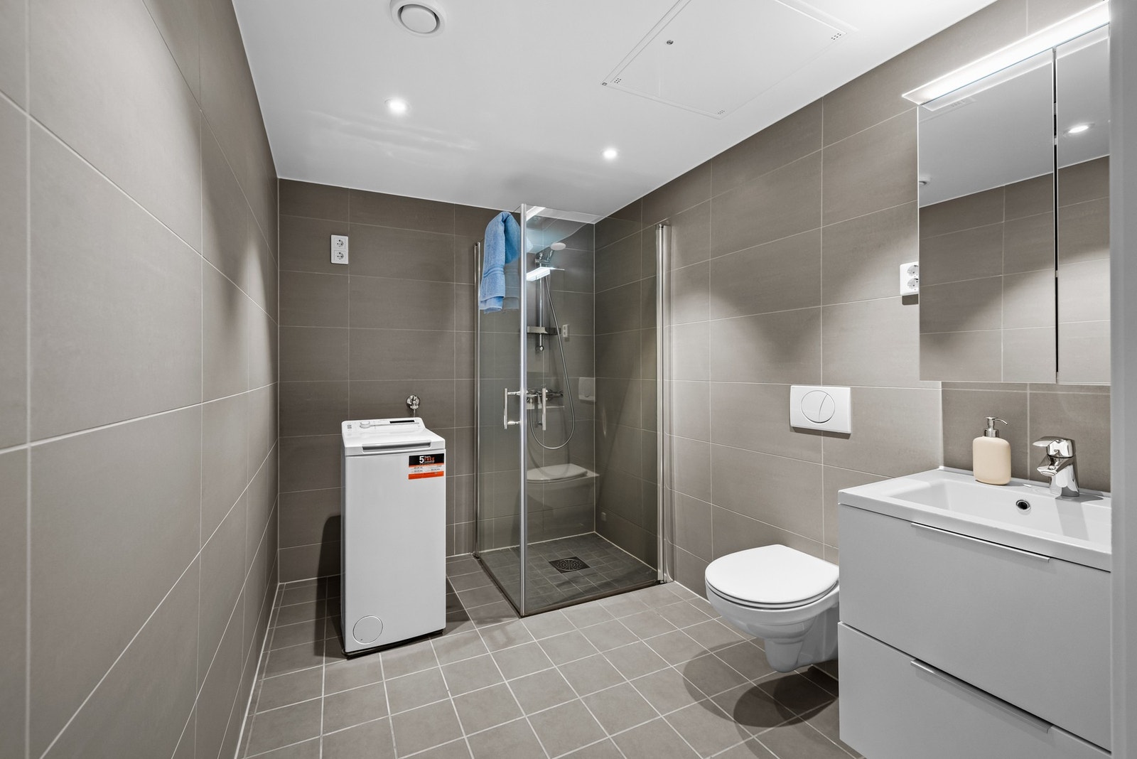 Badet har dusjhjørne med innfellbare dusjdører i glass og armatur tilknyttet hånddusj, vegghengt toalett og opplegg for vaskemaskin og tørketrommel. Galleribilde