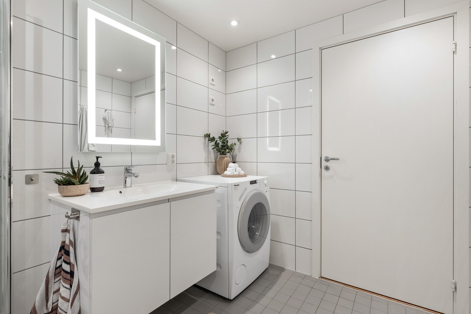 Badet er innredet med vegghengt servantinnredning med ovenpåliggende servant og armatur, speil med over- og sidelys og opplegg for vaskemaskin Galleribilde