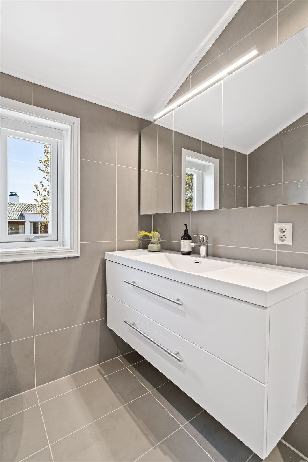 Badet er utstyrt med vegghengt toalett, dusjhjørne med svingbare glassdører, samt baderomsinnredning med servant og speilskap. Galleribilde