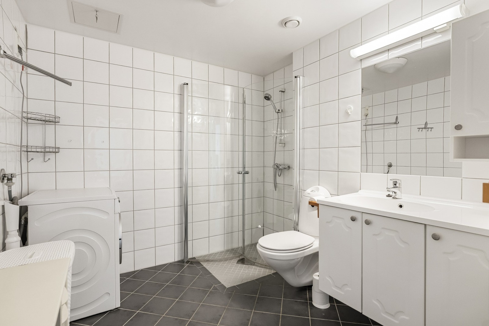 Romslig og praktisk bad med flislagte overflater og en funksjonell utforming som gir god plass til både vaskemaskin og oppbevaring. Galleribilde