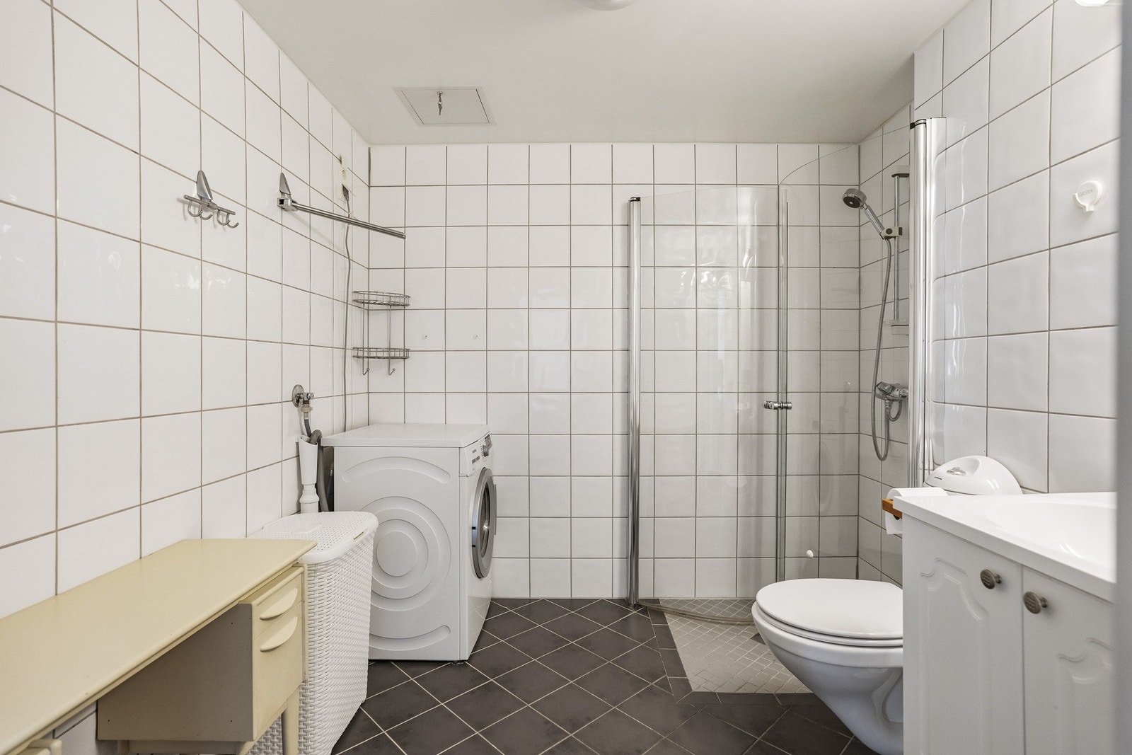 Badet er innredet med dusjløsning med glassvegger, vegghengt toalett og servant med innredning. Lyse fliser på veggene kombinert med mørkt gulv gir et tidløst og stilrent uttrykk Galleribilde