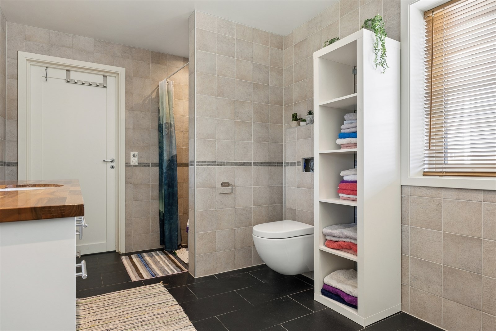 Badet har flislagte gulv og vegger og malt panel i himling. Rommet har vegghengt toalett, dusjnisje med dusjgarnityr på vegg, og innredning med servant i vegghengt skap. Det er sentralt avtrekk med tilluft fra tilstøtende rom. Galleribilde