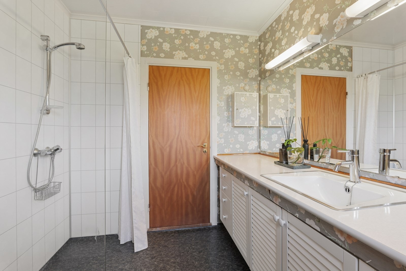 Rommet har dusjvegg og en lang benkeplate med nedfelt servant og rikelig med oppbevaringsplass. Rommet har et renoveringsbehov. Galleribilde