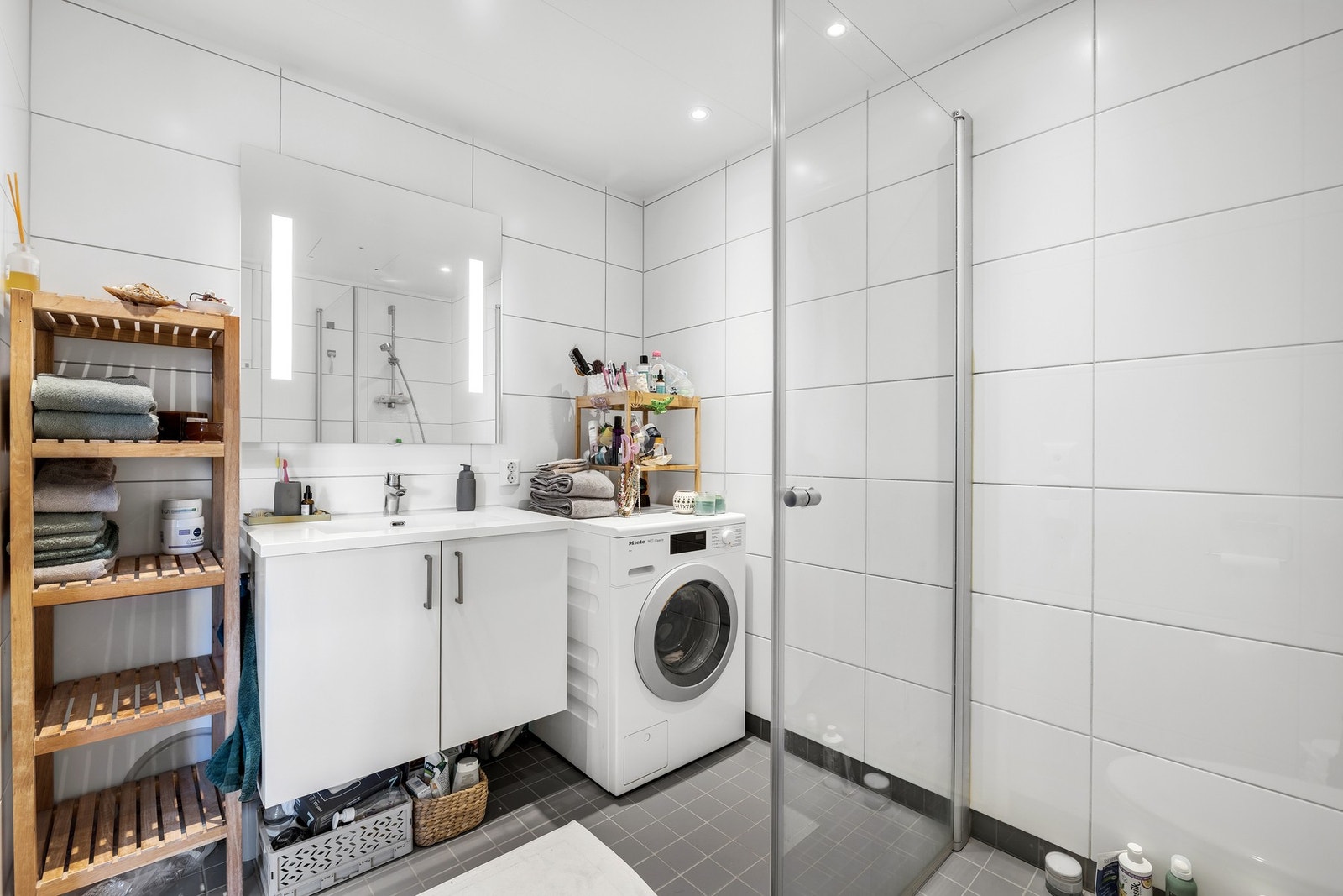 Badet inneholder innfellbare og plassbesparende dusjdører i glass, dusjbatteri og garnityr montert på vegg, kran og avløp for vaskemaskin og veggmontert WC. Galleribilde