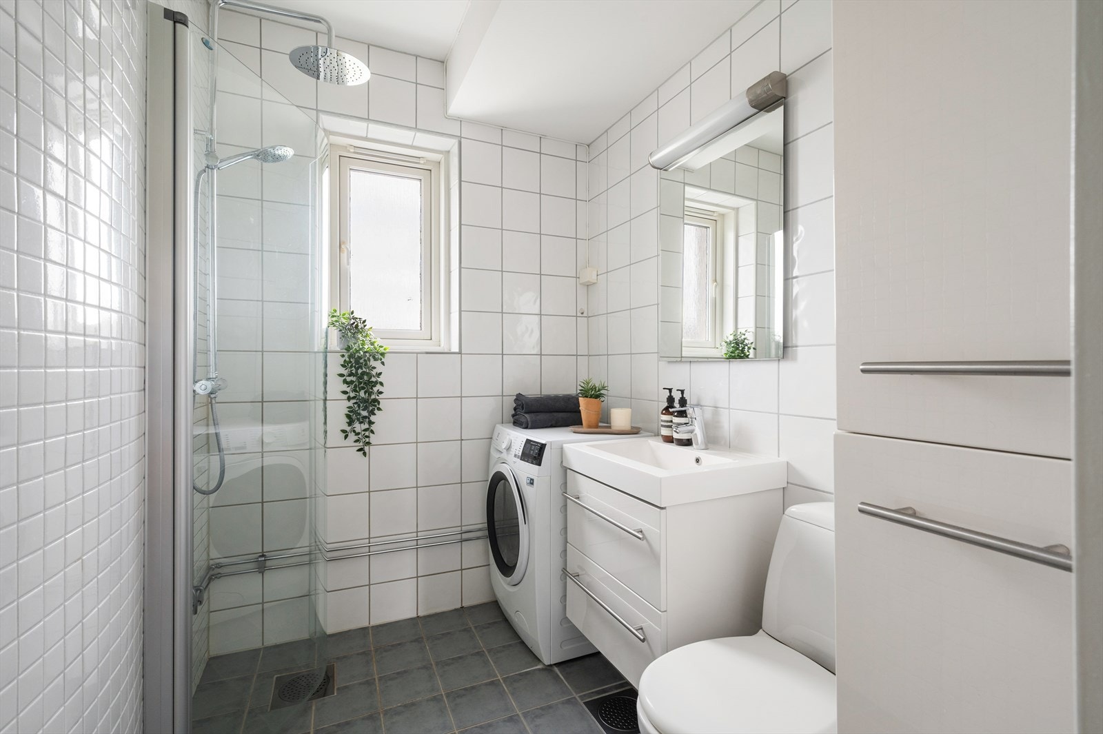 Tidløst flislagt bad med varmekabler i gulv. Badet har gulvmontert wc, dusj på gulv med dører i herdet glass, servant med hvit skuffeseksjon, skap for oppbevaring, samt speil med belysning. Opplegg for vaskemaskin. Galleribilde