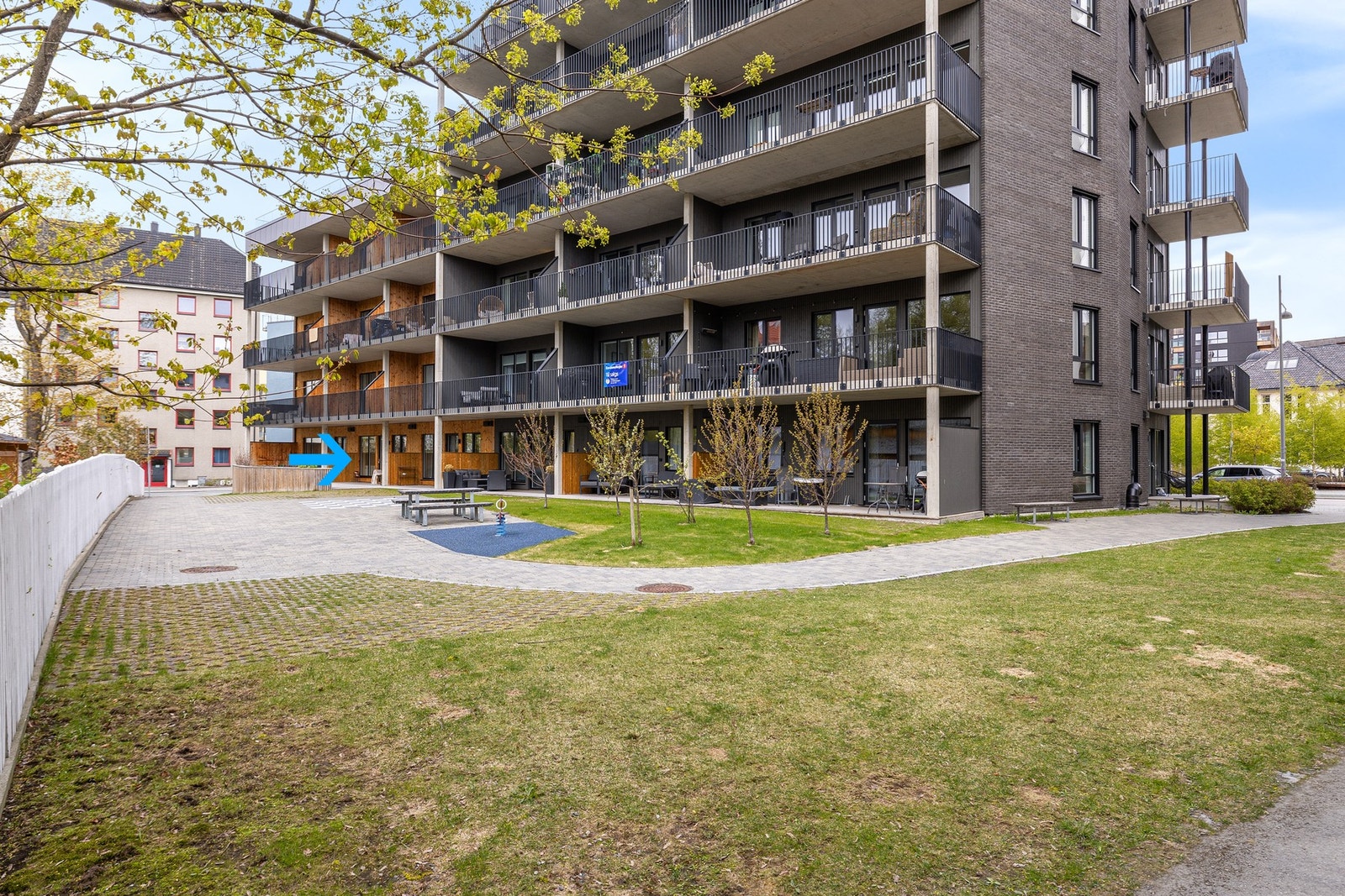 Fasadebilde. Leiligheten er markert med blå pil. På den sørlige delen av bygget har du et pent opparbeidet felles uteareal. Oppleves grønt og frodig på sommerhalvåret. Galleribilde