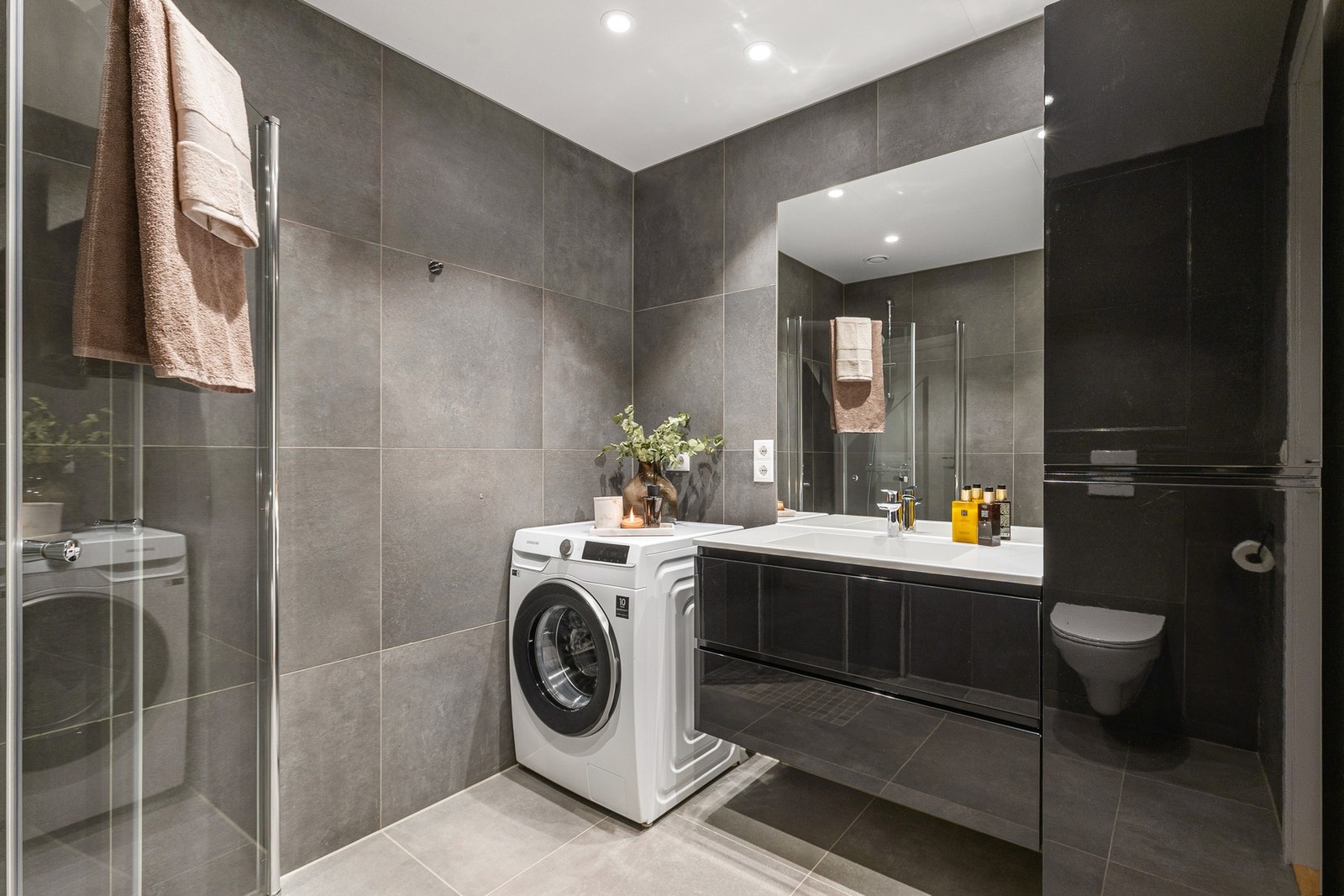 Badet inneholder innfellbare og plassbesparende dusjdører i glass, dusjbatteri og garnityr montert på vegg, veggmontert WC og kran og avløp til vaskemaskin. Spotter i himling. Galleribilde