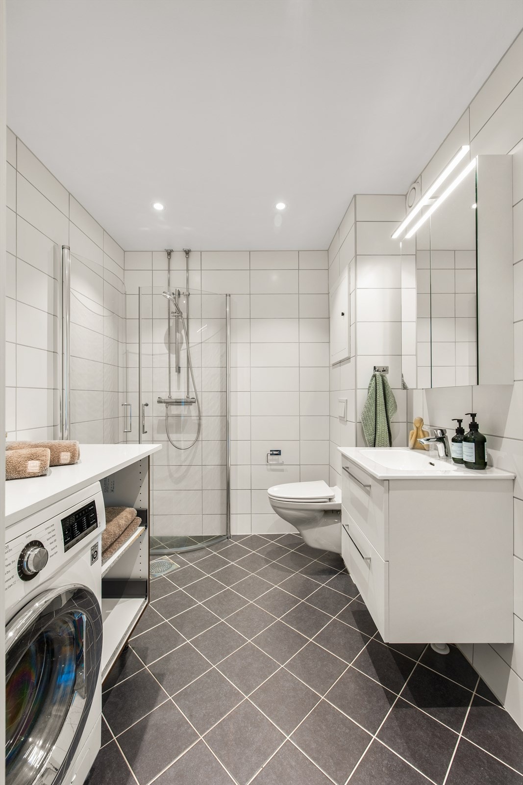 Delikat flislagt baderom (2021) med varmekabler i gulv og downlights i himling. Rommet inneholder bl. a. veggmontert toalett og opplegg for vaskemaskin. Galleribilde