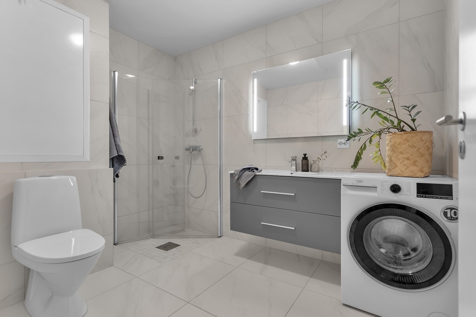 Badet er meget delikat og helfliset, med et moderne design som kombinerer funksjonalitet og eleganse. Dusjnisjen har plassbesparende glassvegger, og mosaikkfliser i dusjsonen tilfører et eksklusivt preg. Galleribilde