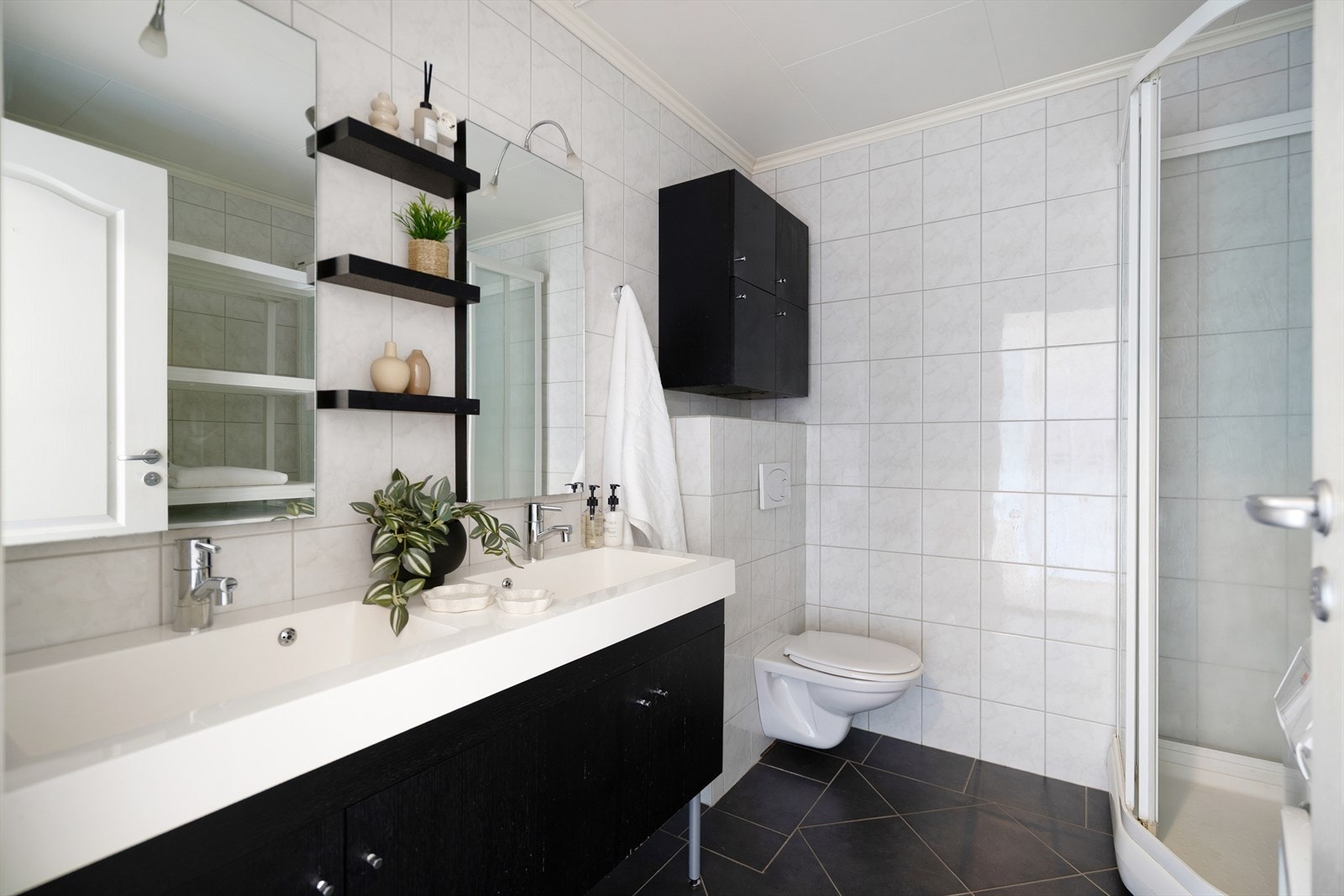 Badet har flislagt gulv men varmekabler, vegghengt toalett, dobbel servant med oppbevaring og dusjkabinett. Galleribilde