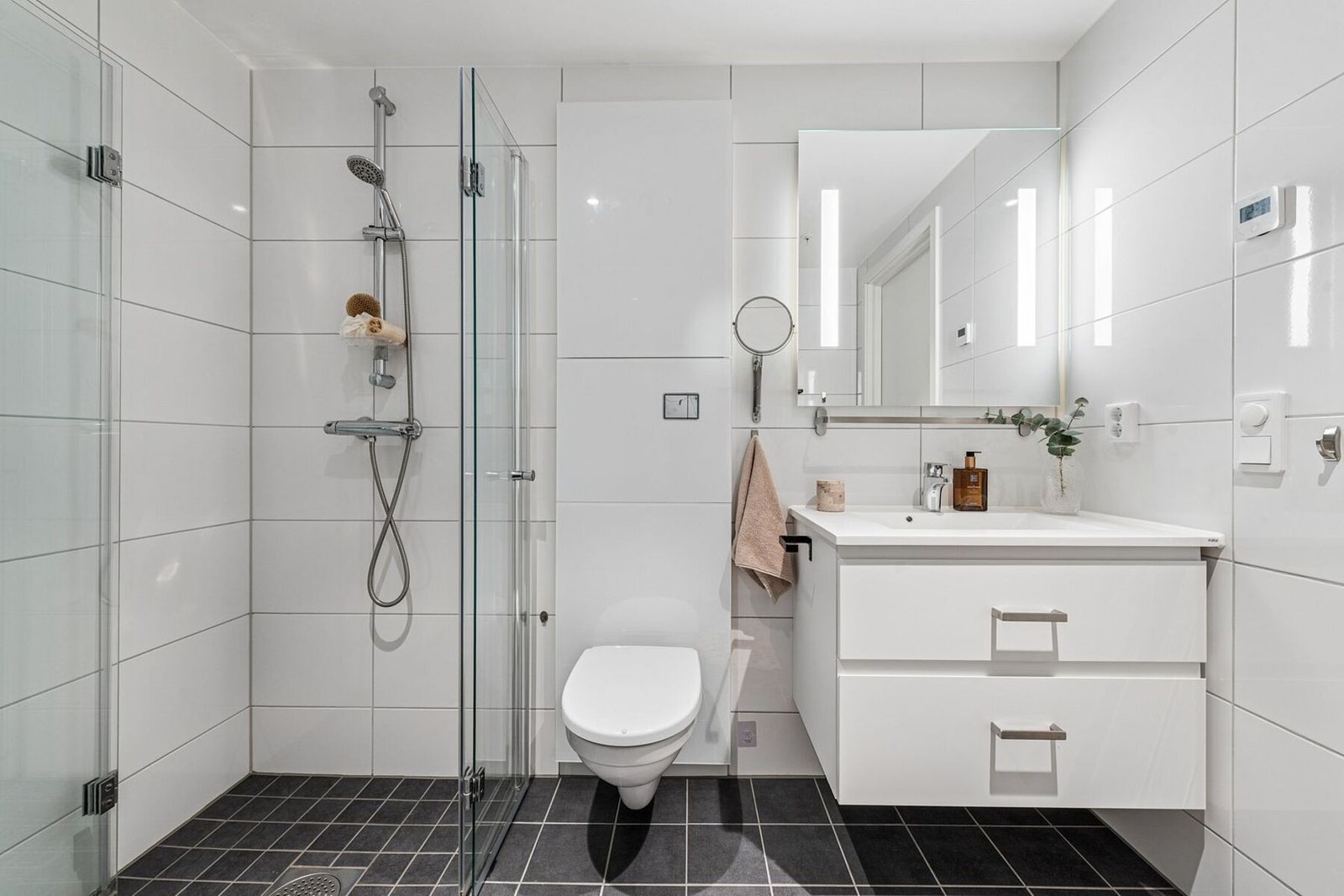 Badet har dusjsone med vendbare glassdører i herdet glass. Romslig baderomsinnredning med brede skuffer og heldekkende servant, samt speil med integrert belysning. Galleribilde